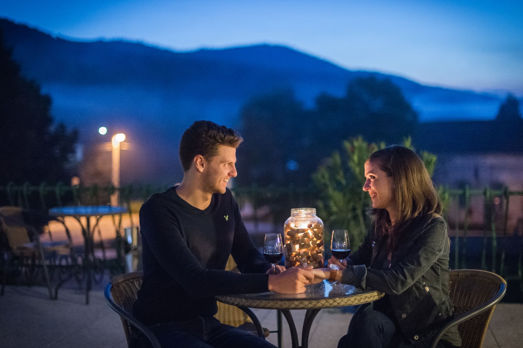 Egy fiatal pár ül a teraszon hangulatos fények mellett este és vörösbort isznak