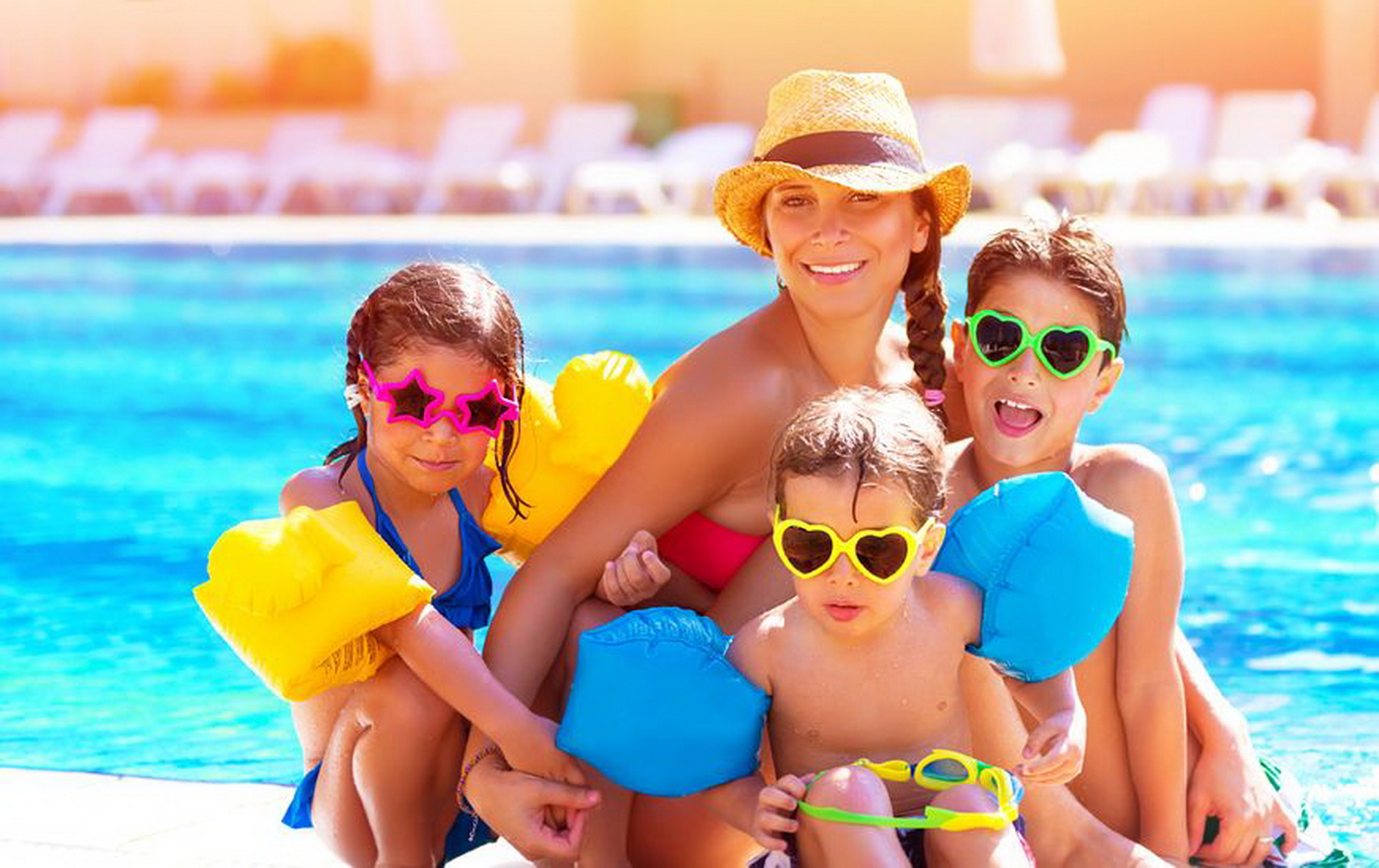 Két kisfiú, egy lány nagyszemüvegben az anyjukkal a medence partján a napsütésben