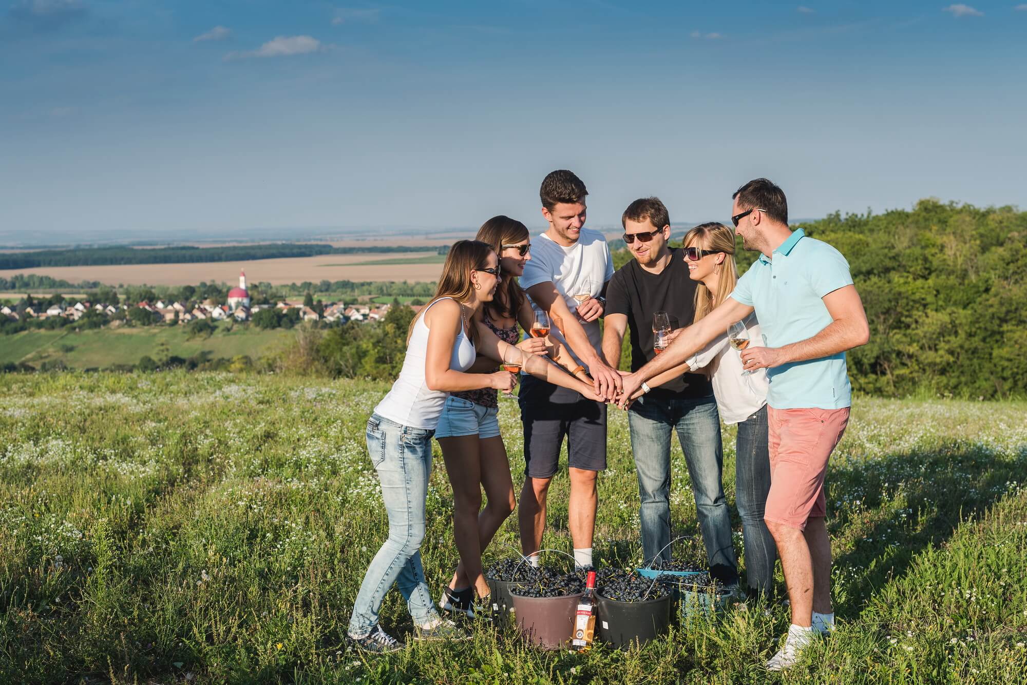 Egy csapat fiatal kezüket összerakva mosolyognak a napsütésben borospohárral a kezükben a Fekete-hegyen, lábuk előtt szőlők vödörben