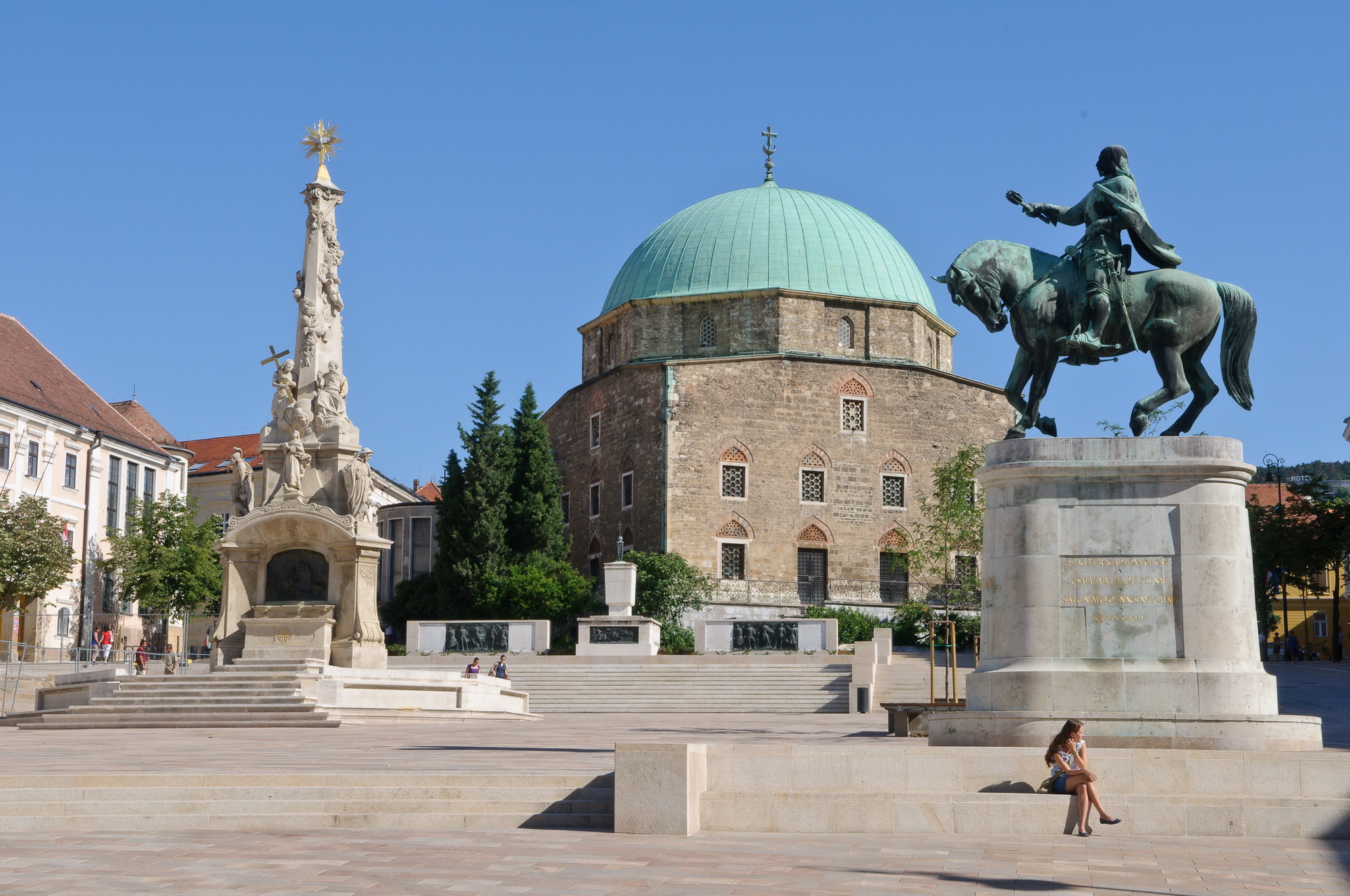 A dzsámi Pécs főterén, előtérben Hunyadi János lovasszobra és a Szentháromság-szobor