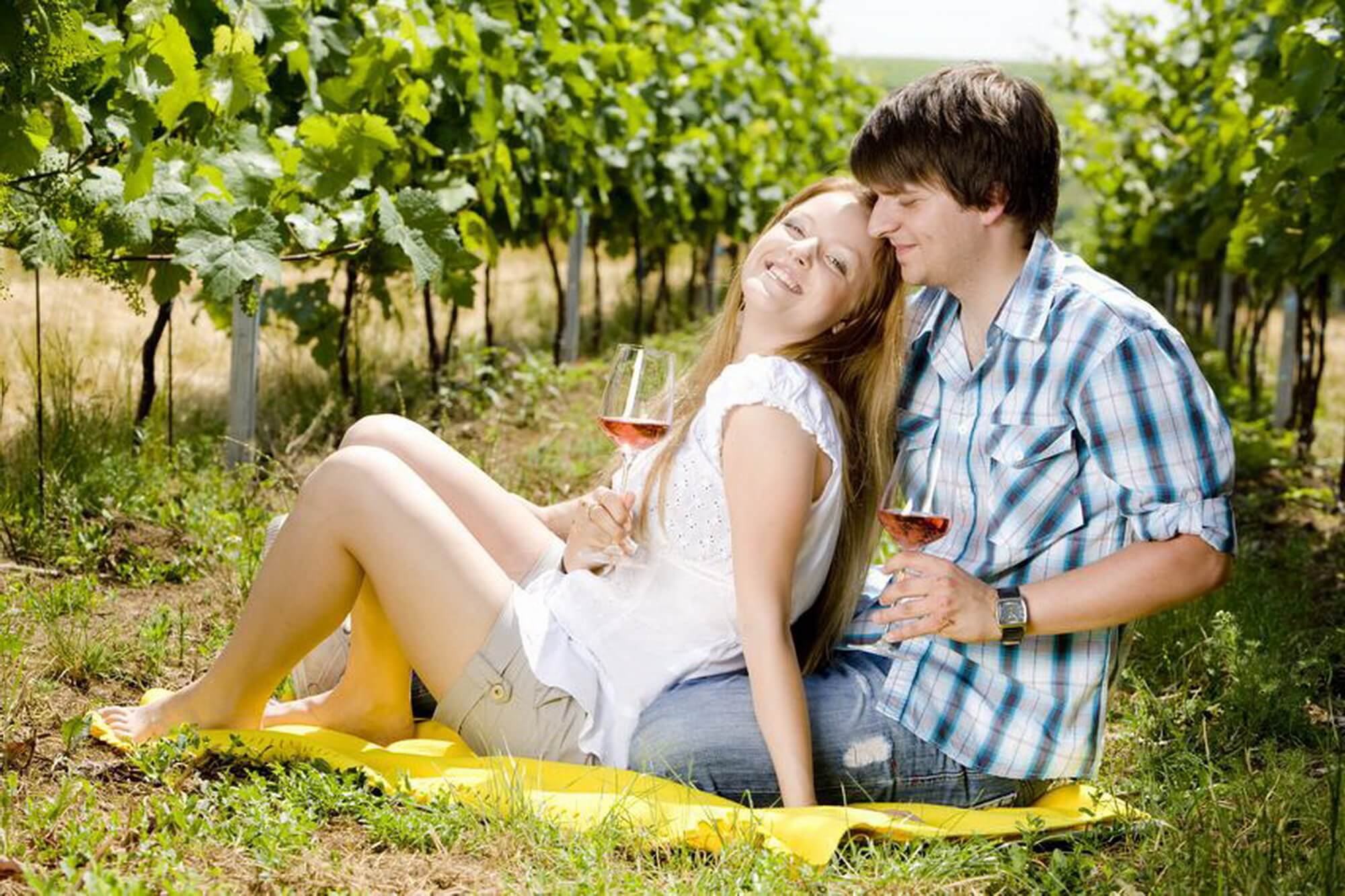 Mosolygó fiatal pár bort kortyolgat a szőlőtőkék között a napsütésben